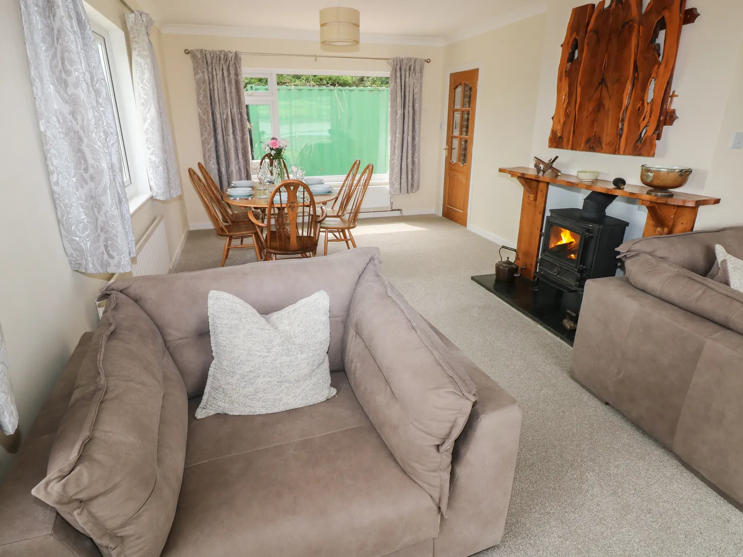 Behind a large chair is a dining table and wood burner. There are also two large windows.
