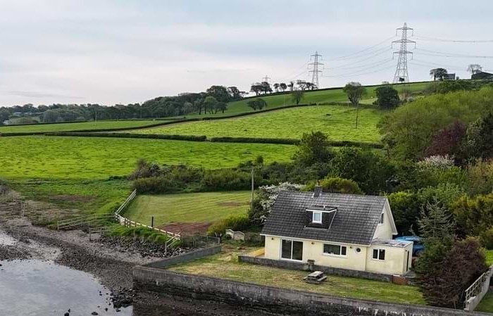A view from the river showing shearwater cottage's riverside garden. Behind the cottage are green rolling fields with trees on the horizon.